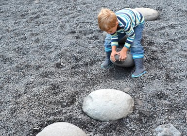 Ein kleiner Junge in blauen Gummistiefeln spielt auf einem Kieselstrand und balanciert auf einem großen Stein.