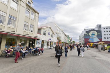 Eine belebte Straße in Akureyri, mit Menschen, die an Cafés sitzen und Passanten, die vorbeigehen, umgeben von bunten Gebäuden.