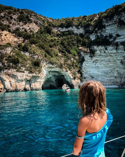 Ein Kind in einem blauen Badeanzug steht am Boot und blickt auf das glitzernde Wasser der blauen Höhlen von Paxos.