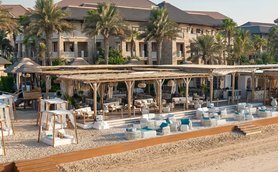 Ein luxuriöser Strandbereich mit gemütlichen Liegen und einem stilvollen Restaurant im Hintergrund, umgeben von Palmen.