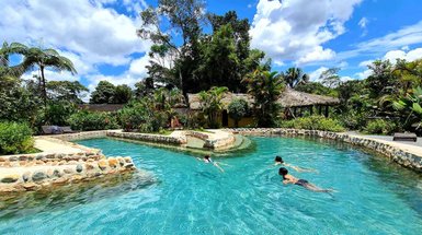 Drei Kinder schwimmen fröhlich in einem klaren Pool, umgeben von üppigem Grün und strahlend blauem Himmel.