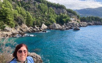 Eine Person mit Brille lächelt vor einer malerischen Küstenlandschaft mit klarem Wasser und grünen Hügeln.