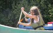 Ein Mädchen mit langen Haaren paddelt energisch in einem Kanu, während ein weiterer Paddler im Hintergrund zu sehen ist.