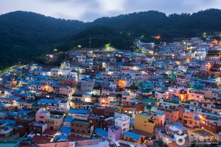 Die bunten Häuser des Gamcheon Culture Village leuchten in der Dämmerung, umgeben von sanften Hügeln und einem klaren Himmel.