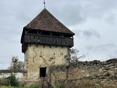 Ein alter, verfallener Turm mit einem schrägen Dach steht inmitten von hohem Gras und umgeben von einer steinernen Mauer.
