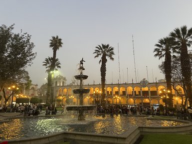 Eine belebte Plaza mit einer großen Brunnenanlage, umgeben von Palmen und historischen Gebäuden, die abends beleuchtet sind.