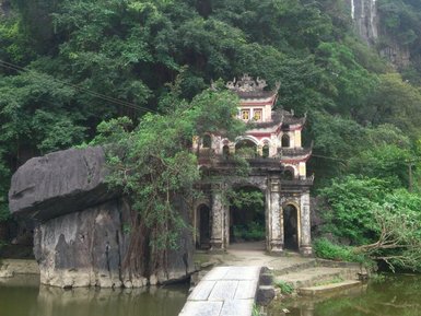 Ein malerischer Tempel mit bunten Verzierungen steht am Ufer eines ruhigen Teichs, umgeben von üppigem Grün und Felsen.