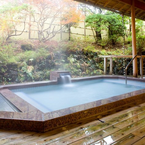 Ein elegantes, dampfendes Onsen mit einem Wasserfall, umgeben von üppigem Grün und Holzverkleidung, strahlt Ruhe aus.