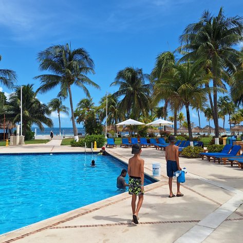Zwei Jungen in Badehosen gehen am Pool entlang, umgeben von Palmen und Liegen unter einem strahlend blauen Himmel.