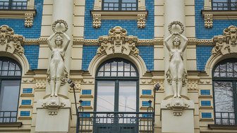 Zwei elegante Statuen in Jugendstil, die mit Kränzen auf den Händen die Fassade eines Gebäudes zieren.