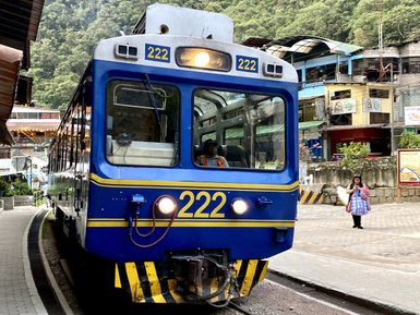Ein blauer Zug mit der Nummer 222 steht an einem Bahnhof, umgeben von üppigen grünen Bergen und einem kleinen Dorf.