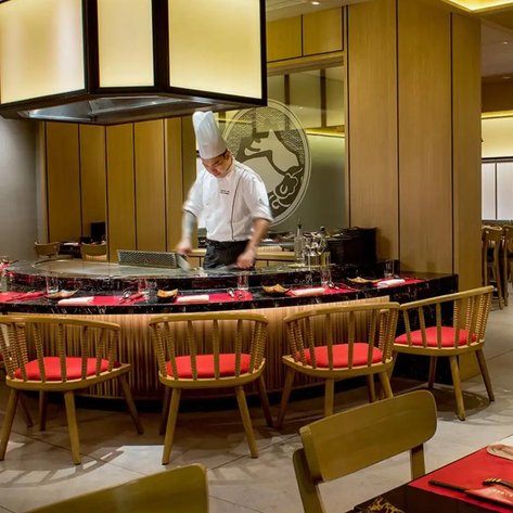 Ein Koch in einem eleganten Restaurant bereitet Speisen an einer zentralen Grillstation vor, umgeben von stilvollen Möbeln.