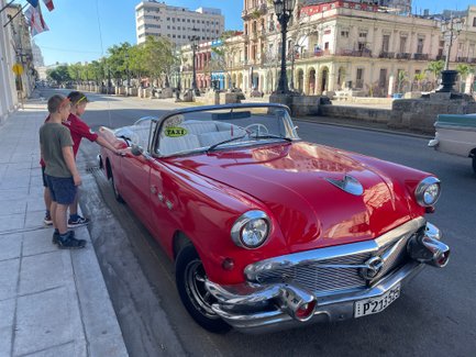 Zwei Jungen stehen neben einem roten Oldtimer-Cabrio, das als Taxi gekennzeichnet ist, auf einer ruhigen Straße.