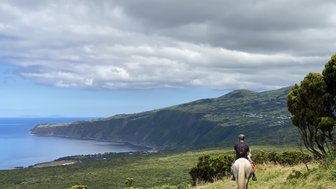 Ein Reiter auf einem weißen Pferd genießt die atemberaubende Aussicht auf die Küste und das grüne Hügelland im Hintergrund.