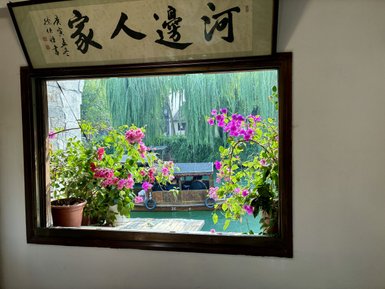 Ein Fensterrahmen mit bunten Blumen, das einen Blick auf einen ruhigen Kanal und ein traditionelles Boot bietet.