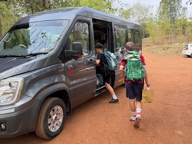 Zwei Kinder steigen in einen grauen Minibus ein, während sie Rucksäcke tragen und ein Kind eine Papiertüte hält.
