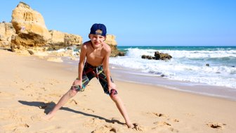 Ein fröhlicher Junge steht am Strand, mit weit gespreizten Beinen und einem breiten Lächeln, während die Wellen sanft an den Strand rollen.