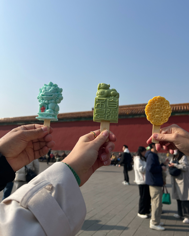 Drei Personen halten farbenfrohe Eis am Stiel in Form von chinesischen Symbolen vor einem historischen Hintergrund.