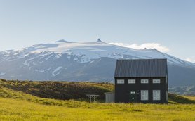 Ein schwarzes Sommerhaus steht in einer grünen Wiese, mit schneebedeckten Bergen im Hintergrund, die unter blauem Himmel leuchten.