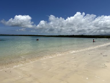 Ein ruhiger Strand mit feinem Sand, wo Kinder im flachen Wasser spielen und große, weiße Wolken am blauen Himmel schweben.