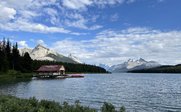 Ein malerischer See mit einem Holzhaus am Ufer, umgeben von majestätischen Bergen und einem klaren blauen Himmel.