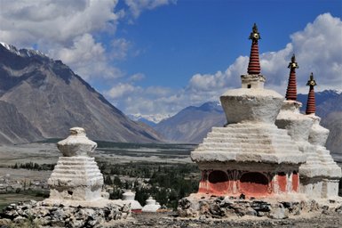 Drei beeindruckende Stupas mit roten Spitzen stehen vor einer majestätischen Berglandschaft unter einem blauen Himmel.