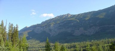 Zwei Personen stehen auf einem Campingplatz mit grünen Zelten, umgeben von hohen Bäumen und majestätischen Bergen im Hintergrund.