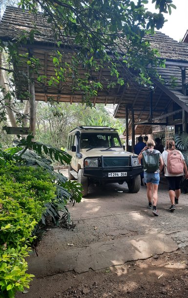 Zwei Personen mit Rucksäcken gehen in Richtung eines überdachten Eingangs, während ein Geländewagen parkt.