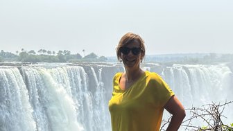 Eine Frau in einem gelben Shirt lächelt vor den beeindruckenden Victoriafällen, umgeben von üppigem Grün.