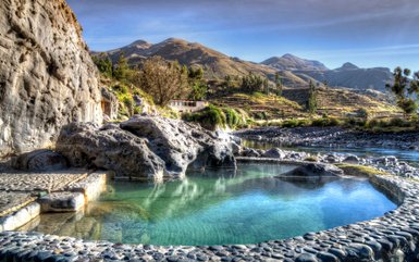 Ein klarer Thermalpool umgeben von Felsen und üppiger Vegetation, mit sanften Hügeln im Hintergrund.