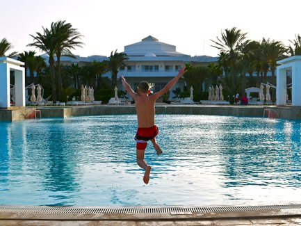 Ein Junge springt fröhlich in einen glitzernden Pool, umgeben von Palmen und einem eleganten Hotel im Hintergrund.