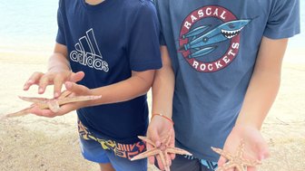 Zwei Jungen stehen am Strand und halten jeweils einen Seestern in der Hand, während das Wasser im Hintergrund glitzert.