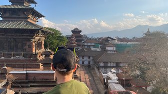 Ein Junge mit einer schwarzen Kappe blickt auf die beeindruckende Architektur von Kathmandu, umgeben von Bergen und Wolken.