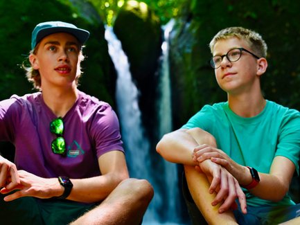 Zwei Jungen sitzen am Ufer eines klaren Flusses, umgeben von üppigem Grün, während ein Wasserfall im Hintergrund plätschert.
