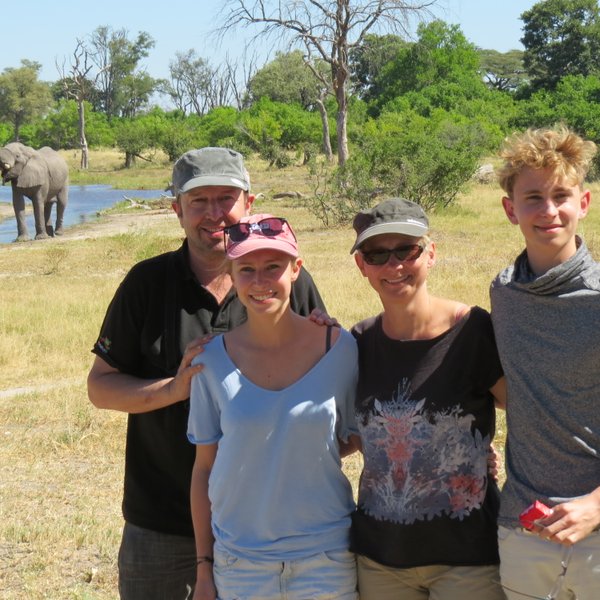 Eine Familie posiert fröhlich vor einem Elefanten, der im Hintergrund an einem Fluss steht, umgeben von üppiger Natur.
