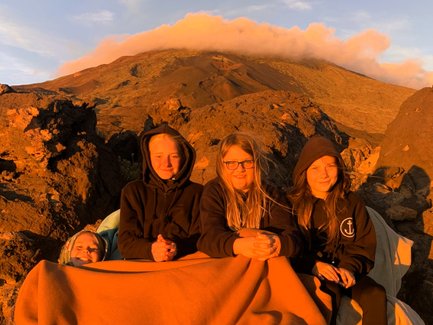 Fünf Jugendliche sitzen auf einer Decke in einer vulkanischen Landschaft, umgeben von beeindruckenden Felsen und warmem Licht.