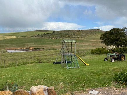 Eine weitläufige Wiese mit einem Spielplatz und einem alten Traktor, umgeben von sanften Hügeln und bewölktem Himmel.