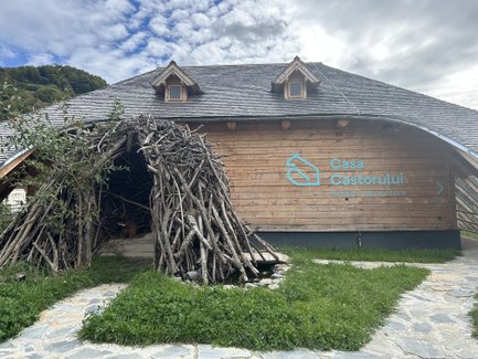 Ein Holzgebäude mit einem großen, kunstvoll gestalteten Eingang aus Ästen, umgeben von grünem Gras und einem gepflasterten Weg.