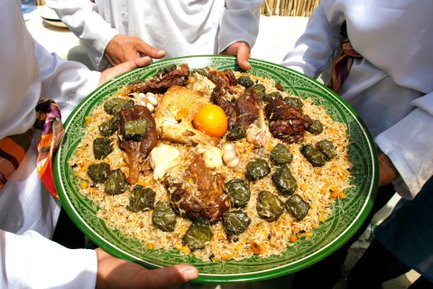 Ein großer, grüner Teller mit duftendem Plov, garniert mit Hähnchen, Lamm und gefüllten Weinblättern, wird von mehreren Händen gehalten.