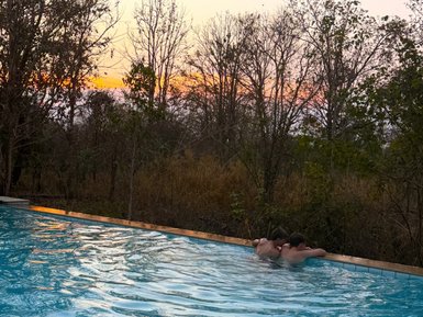 Zwei Personen entspannen im Pool, umgeben von barem Baumkleid und goldenen Sonnenstrahlen am Horizont.