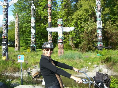 Eine fröhliche Radfahrerin posiert vor bunten Totempfählen im Stanley Park, umgeben von üppigem Grün und Sonnenlicht.