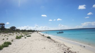Ein sonniger Strand mit feinem, hellem Sand erstreckt sich entlang des türkisfarbenen Wassers, während Menschen im Hintergrund entspannen.