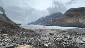 Eine weite, graue Gletscherlandschaft mit steilen Bergen und einer Gruppe von Menschen, die den Gletscher erkunden.