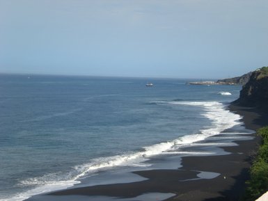 Eine ruhige Küstenlandschaft mit schwarzem Sand, Wellen, die sanft an den Strand rollen, und einem malerischen Boot auf dem Wasser.