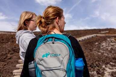 Eine Frau mit einem blauen Rucksack steht auf einem Wanderweg, während eine andere Person im Hintergrund zu sehen ist.