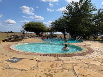 Ein Pool in der Natur mit mehreren Personen, die sich entspannen und schwimmen, umgeben von Bäumen und einem weiten Feld.