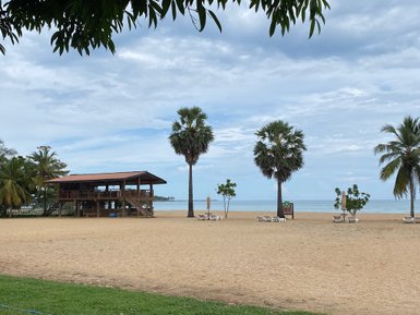 Ein ruhiger Strand mit feinem Sand, umgeben von Palmen und einem Holzgebäude im Hintergrund, unter einem bewölkten Himmel.