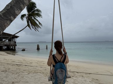 Eine person schwingt entspannt auf einer Holzschaukel am Strand, umgeben von Palmen und feinem Sand.