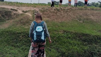 Eine Person mit einem Rucksack geht über eine Holzbrücke, während andere Menschen im Hintergrund stehen und zuschauen.