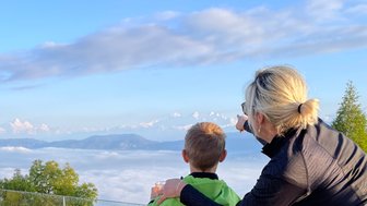Eine Frau zeigt einem Jungen die schneebedeckten Berge, während sie über eine Wolkendecke blicken, die die Landschaft verdeckt.